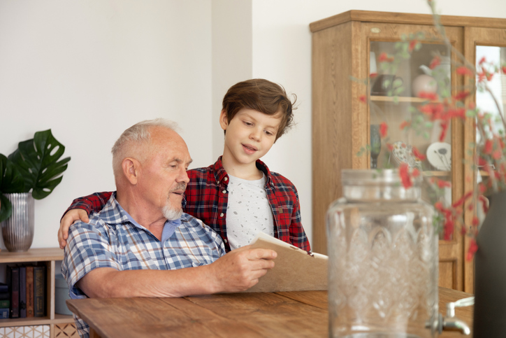 Close up of grandfather spend free time at cozy home kitchen enjoying hobbies together his grandson. Elderly man reading book with cute schoolboy. Lifestyle of a pensioner