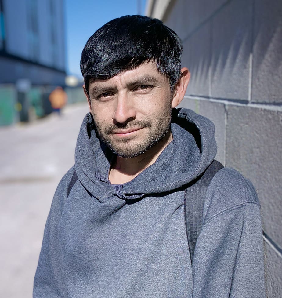 A man with short dark hair and a beard, wearing a gray hoodie and backpack, stands outdoors next to a concrete wall on a sunny day.