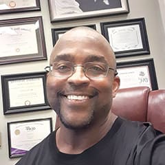 marc A person wearing glasses and a black shirt smiles while seated in a chair, with multiple certificates framed on the wall behind.