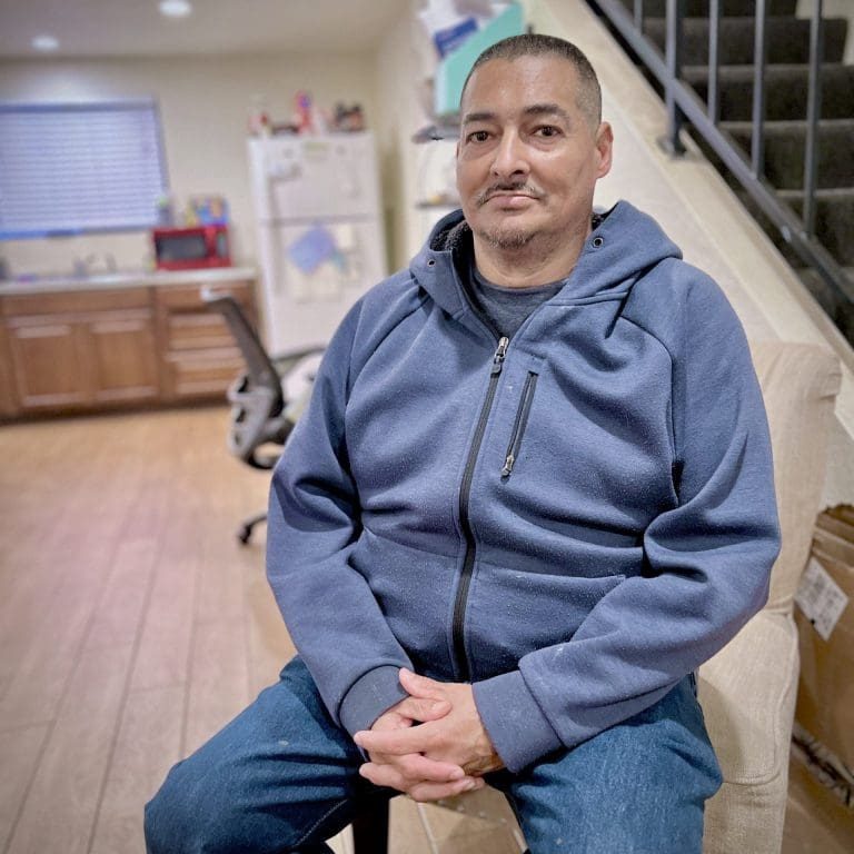 A man in a blue hoodie and jeans sits on a chair indoors, with a kitchen and staircase in the background, perhaps pausing to reflect on his recovery journey or deciding to seek help for addiction.