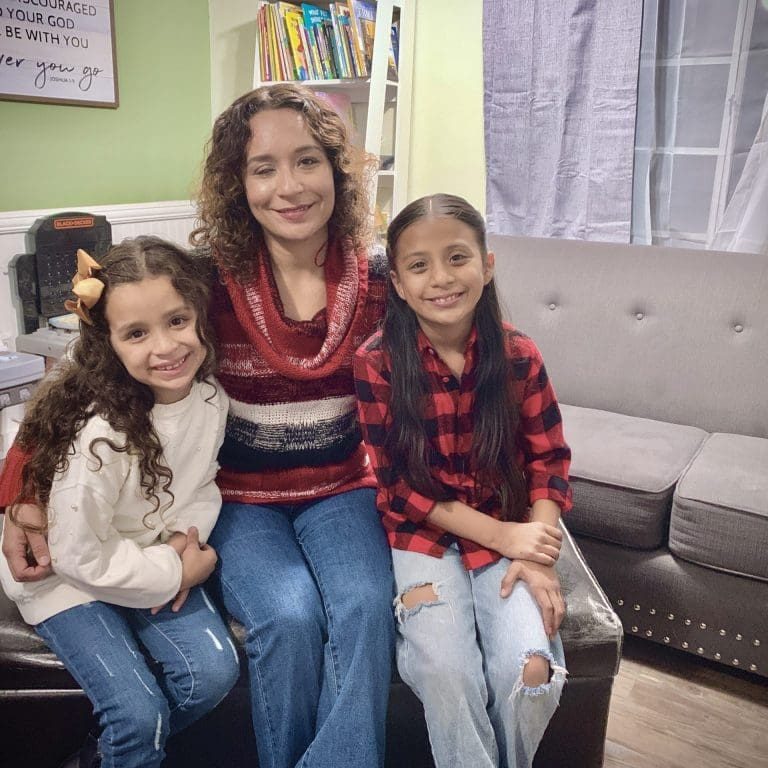 A woman sits on a bench with two young girls beside her, all smiling. In the cozy living room, they enjoy a moment to be still together, surrounded by a gray couch, bookshelf, and wall decor in the background.