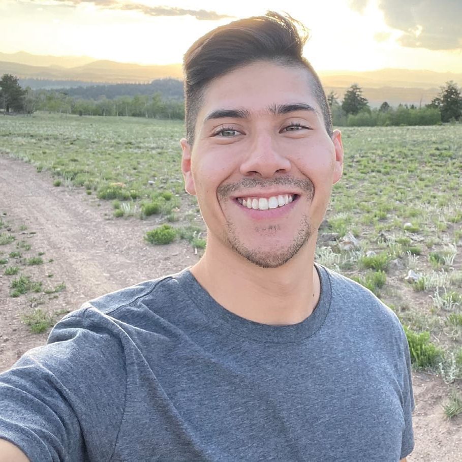 A person in a gray t-shirt smiles while taking a selfie in an open field, capturing the essence of sunset with distant hills behind them, reminiscent of Chef Gs tranquil retreats.