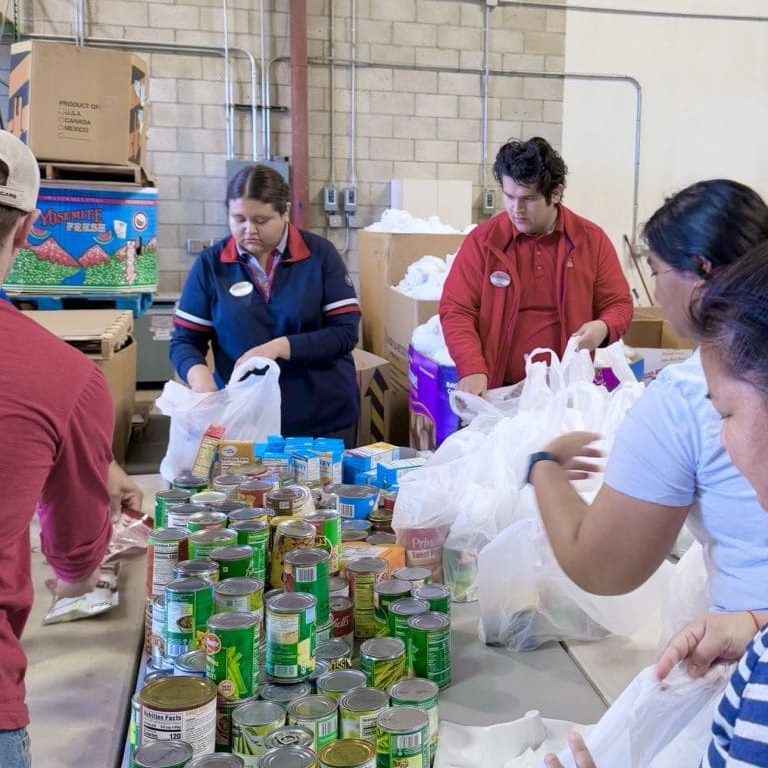 Volunteers participate in Serve Day, packing canned goods and other food items into bags on a long table in a warehouse setting.