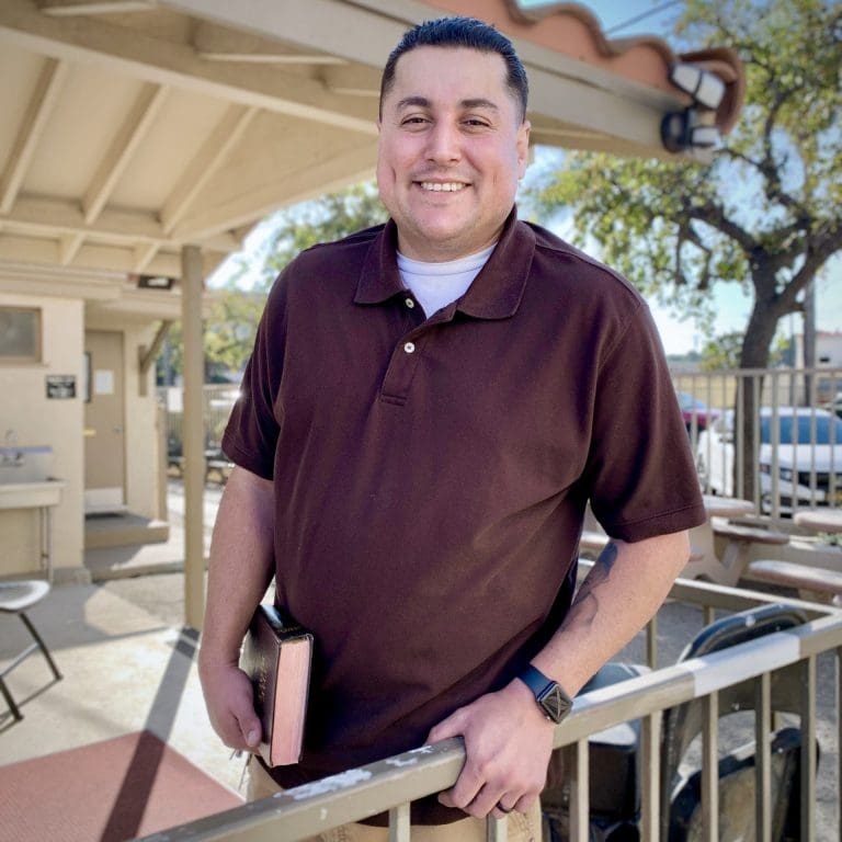 A man in a maroon polo shirt stands outdoors by a railing, holding a book and smiling at the camera, embodying the spirit of Mission and embracing open doors to new opportunities.