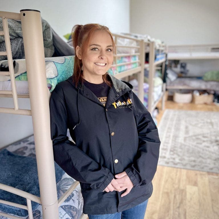 A woman with red hair stands smiling in front of bunk beds in a room with wooden floors, wearing a black jacket and jeans, radiating acceptance as she embraces her true self.