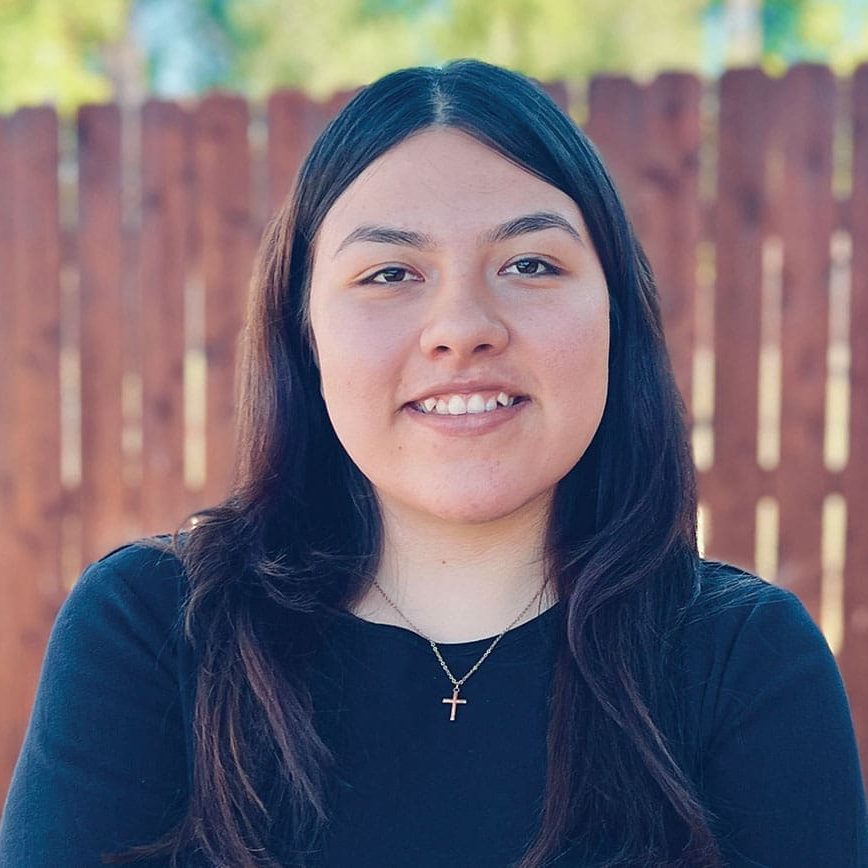 Jocelyn, with long dark hair, wearing a black top and a cross necklace, stands in front of a wooden fence outdoors, ready to share her life-changing message.