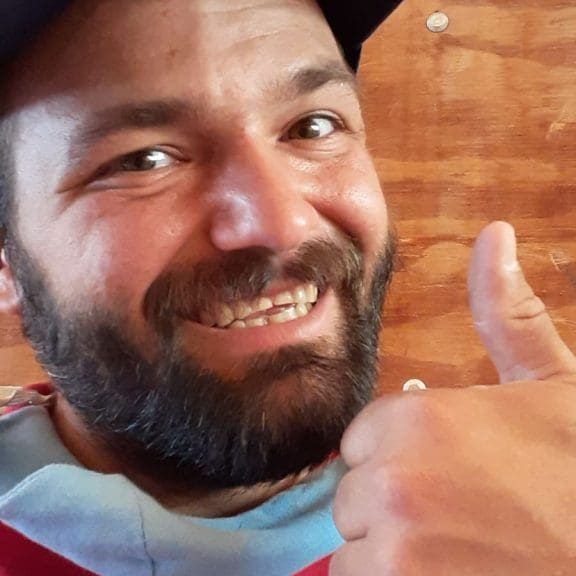 Landon, wearing a beard and a cap, is smiling and giving a thumbs up gesture while standing in front of a wooden wall.