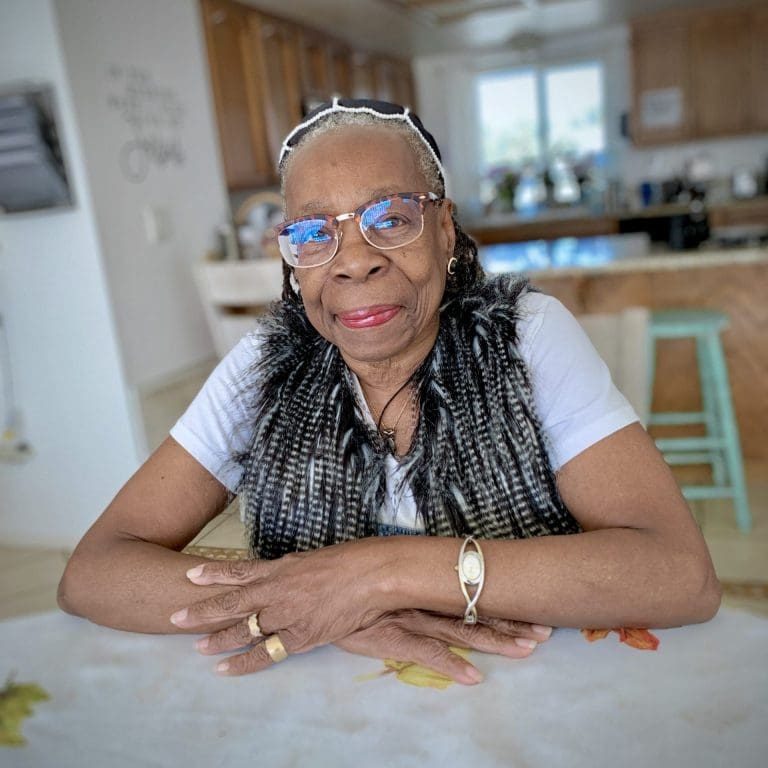 An elderly woman with glasses and a black-and-white vest sits at a table with a fall-themed tablecloth decorated with leaves, drinking tea in a cozy kitchen.