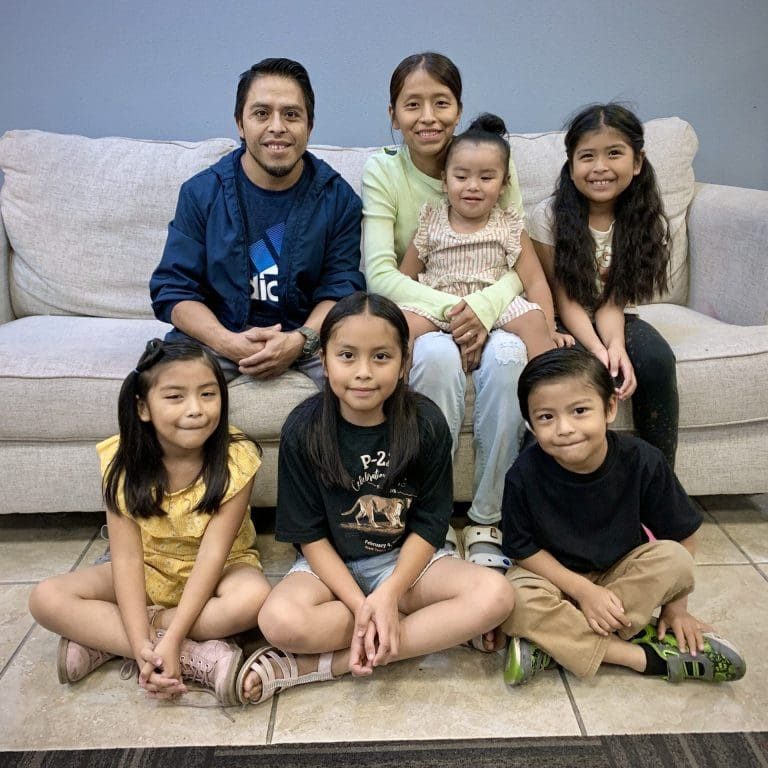 A family of seven, including Felix, two adults, and five children, poses together in a living room, sitting on and in front of a beige couch.