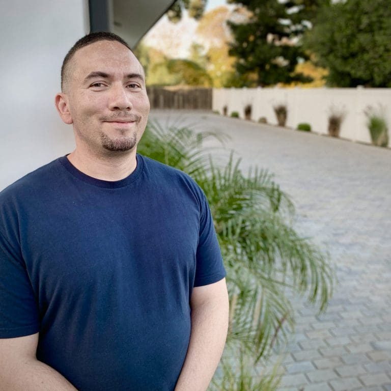 A man in a navy blue t-shirt stands outside on a stone pathway, giving thanks to God for overcoming addiction, with greenery and a white fence in the background.