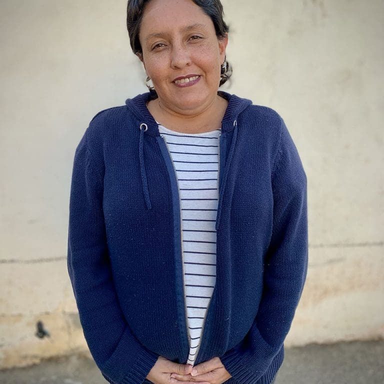 A woman named Teresa with short dark hair, wearing a navy zip-up sweater over a striped shirt, stands in front of a plain light-colored wall, looking at the camera with a slight smile that hints at her quiet courage.