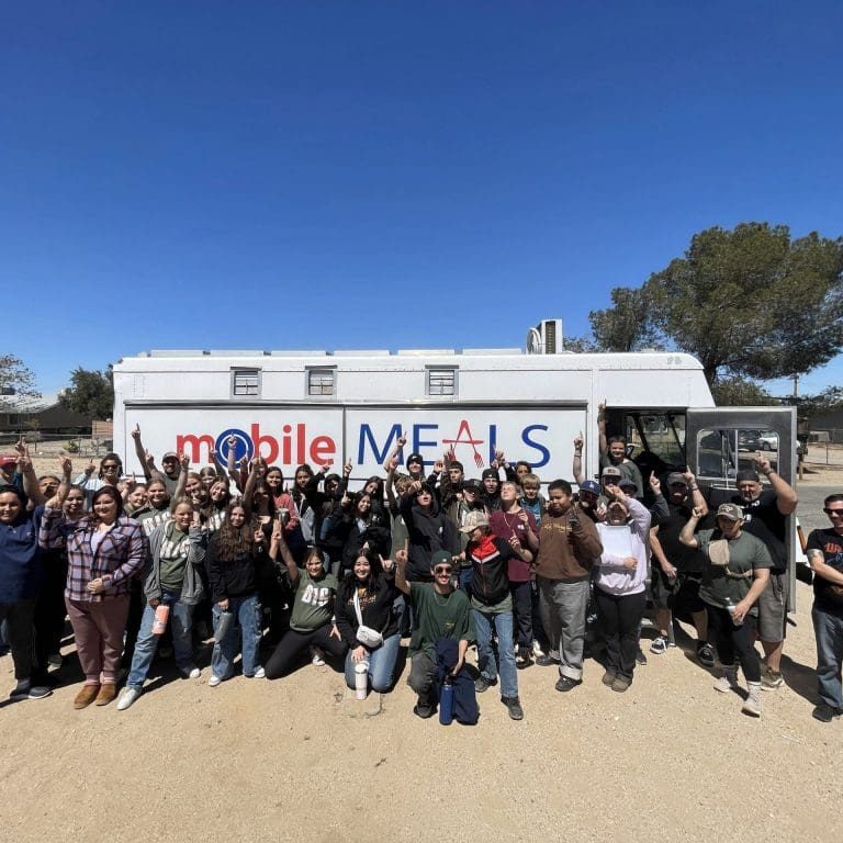 A large group of people stand in front of a white truck labeled Mobile Meals, raising their arms and smiling outdoors on a sunny Big Serve Day, part of the annual event organized by RMA VV.