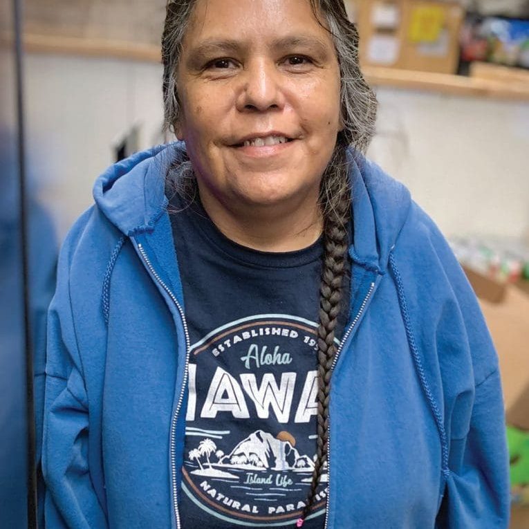 A woman with braided hair wearing a blue hoodie and a Hawaii t-shirt stands indoors, smiling at the camera. Surrounded by boxes and shelves, she radiates hope as she focuses on getting back on your feet after difficult times.