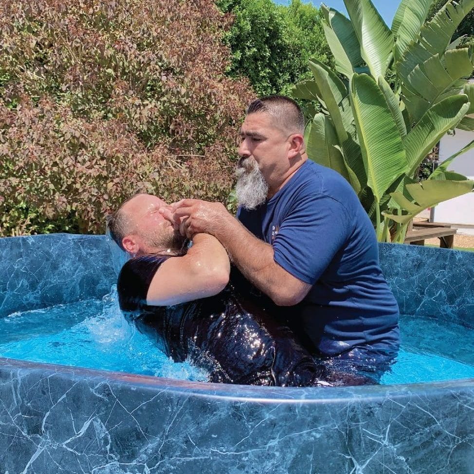 Two men are in a small, round pool; one is submerging the other backward into the water, possibly performing a community baptism. Trees and greenery are visible in the background at RMA SFV.