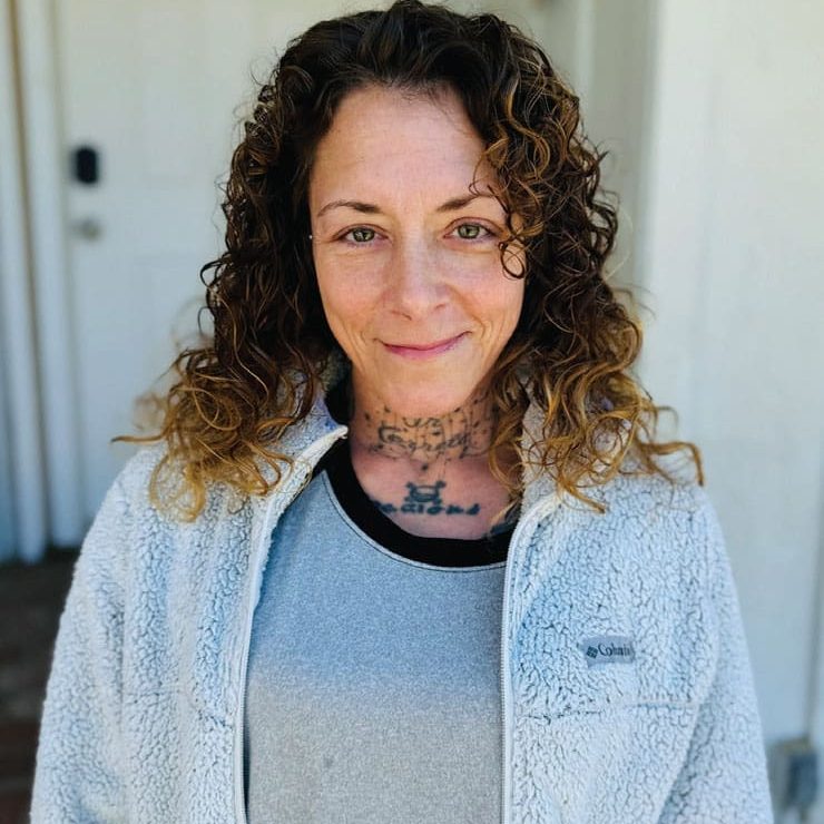 A person with curly hair and tattoos on their neck and chest stands in front of a white door, wearing a gray fleece jacket over a gray shirt, exuding an air of resilience like someone whos been overcoming trauma.