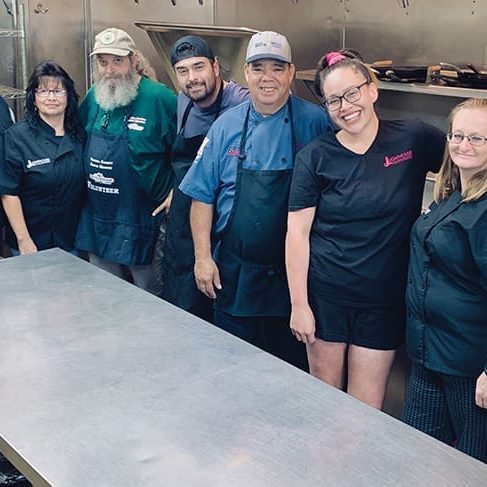 A group of nine people stands together in a commercial kitchen, wearing aprons and smiling at the camera behind a stainless steel table—breaking bread and building futures with RMA VC.