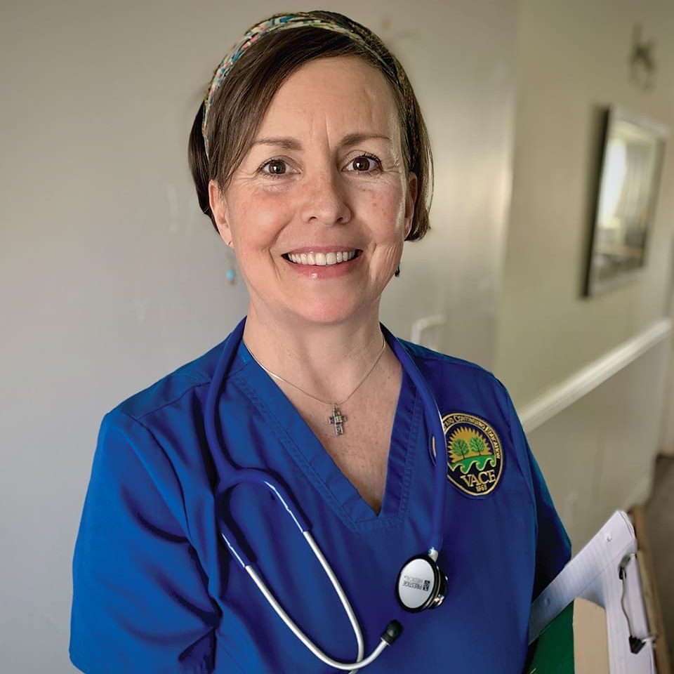 Christina, a woman in blue medical scrubs with a stethoscope and clipboard, smiles at the camera while standing in a hallway, proud to be part of the graduates program.