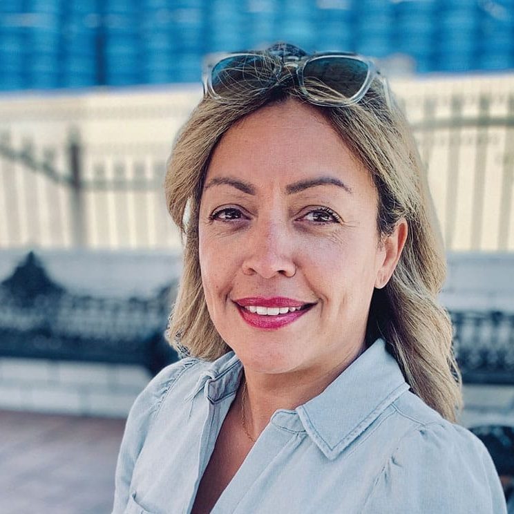 Woman with blonde hair wearing a light blue shirt and sunglasses on her head, smiling outdoors with a bench and fence in the background as she joins the team for a volunteering event.