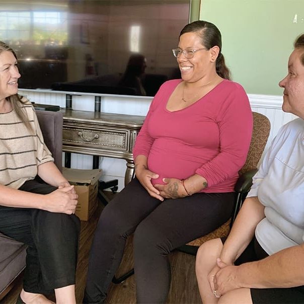 Three women sit in a living room, engaged in conversation. One woman is smiling with her hands on her pregnant belly while the others listen attentively, embodying the warmth and connection highlighted in the Director’s Note.