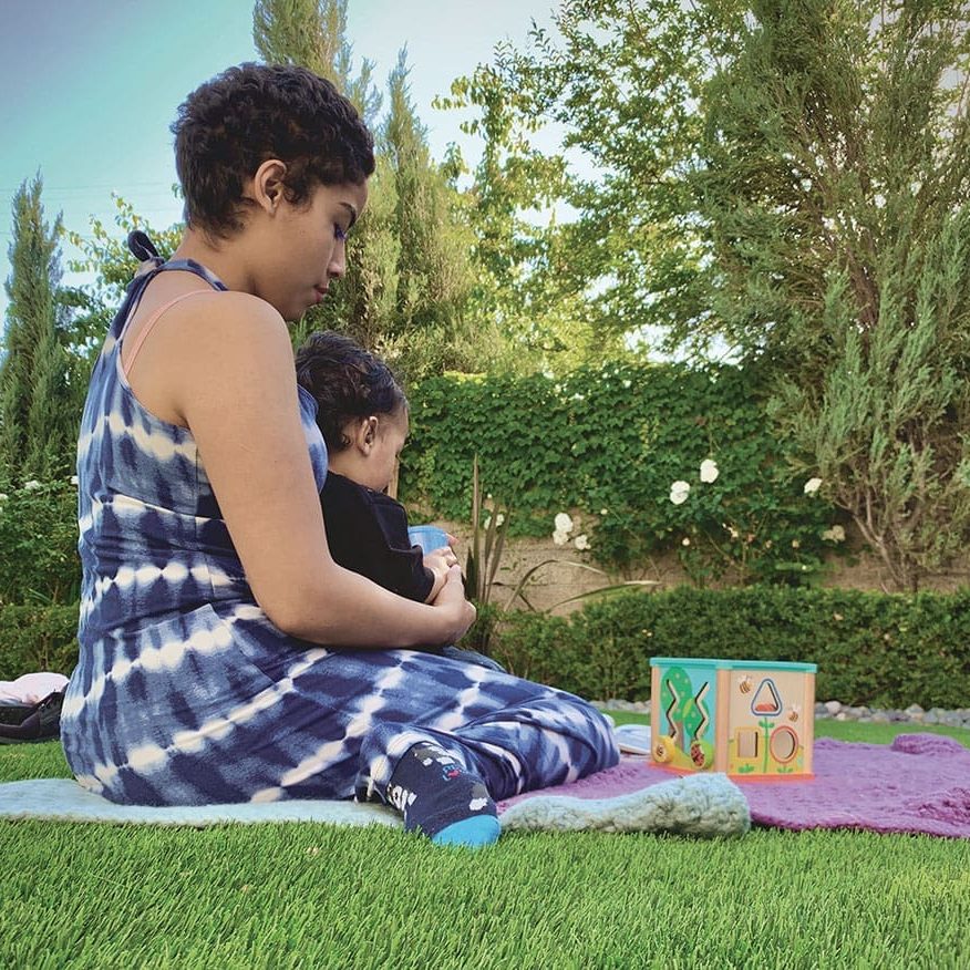 Emily sits on the grass, cradling a baby, with a colorful toy beside them and a green leafy backdrop. Despite having overcome trials, peace surrounds her in this serene moment.