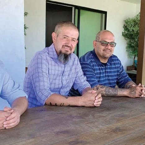 Four men sit at a wooden table outside, all dressed in collared shirts. Their conversation flows from laughter to earnest exchanges as they discuss Felixs journey and the triumphs of addiction recovery, each word carrying the weight of their shared experiences and newfound freedom.