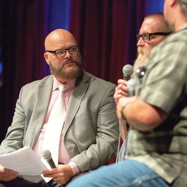 Three men are sitting and holding microphones, with one in a suit holding papers, engaged in a conversation on stage in Victorville with an American flag in the background, discussing a grateful goodbye and reflecting on a lasting legacy.