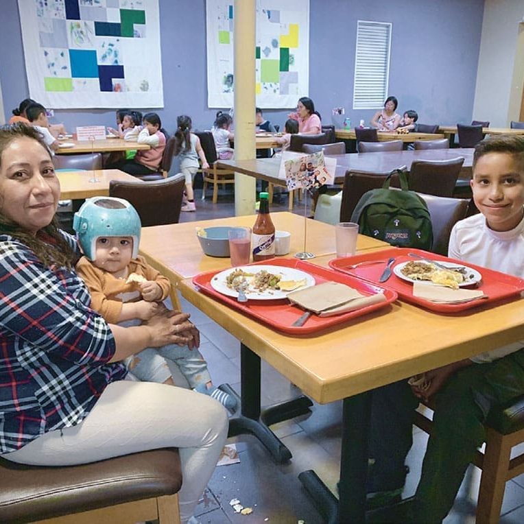 A woman, a baby wearing a helmet, and an older boy sit at a cafeteria table with food trays, sharing a moment of renewed hope while others dine in the background—grateful to feel home again.