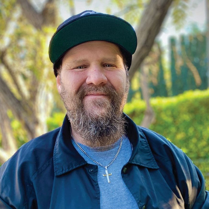 A bearded man named Jason, wearing a black cap, black jacket, and cross necklace, stands outdoors among trees and greenery, embracing a new life.