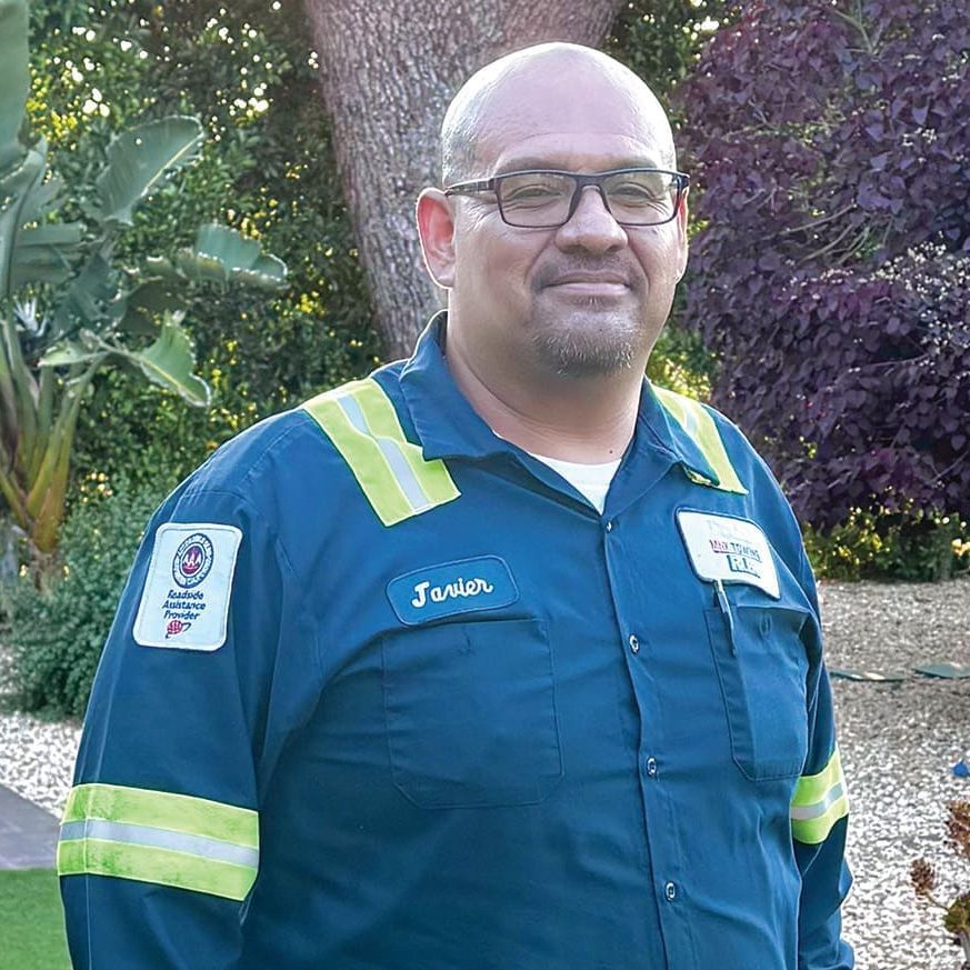 Javier, wearing glasses and a work uniform with reflective stripes, stands outdoors in a garden setting, his expression shaped by hope and experience from over three decades on the job.