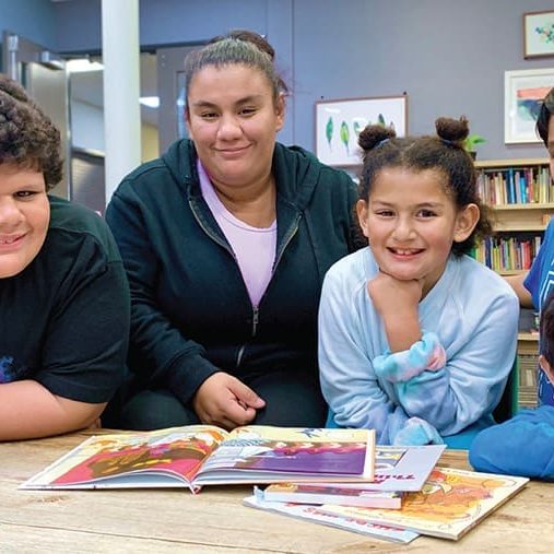 A group of five children are smiling around a table with open books in a bright, cozy room filled with bookshelves, feeling the warmth of being Home Again.