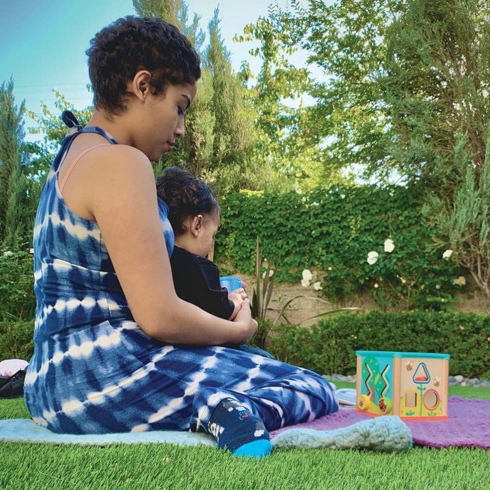Looking back, a woman in a blue tie-dye outfit sits on grass with a child on her lap, next to colorful toy blocks, surrounded by greenery.