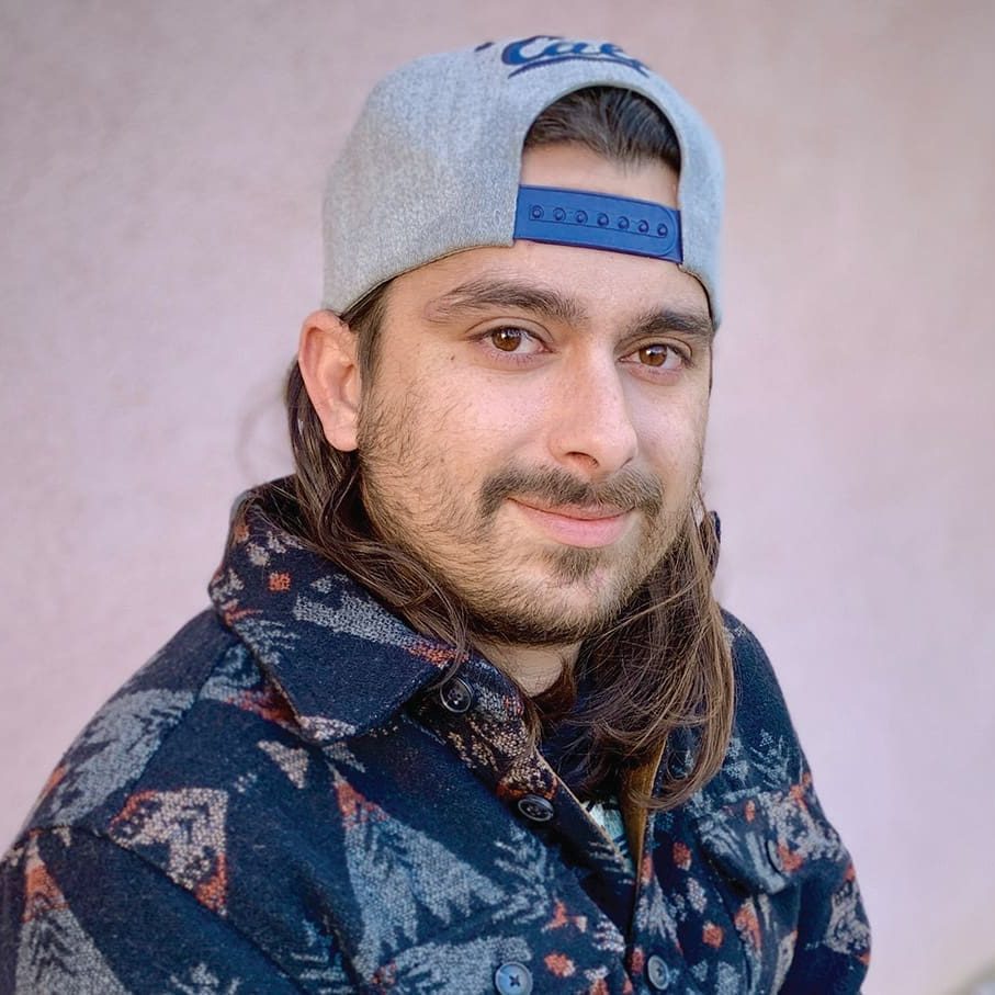 Matthew, a hopeful man with long brown hair and a beard, wears a patterned jacket and a grey baseball cap backwards, posing confidently in front of a plain light-colored background.