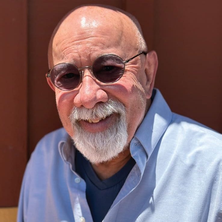 An older man with a bald head, white beard, and sunglasses smiles while wearing a light blue shirt, standing in sunlight before a brown wooden wall—one of the dedicated RMA VC volunteers.