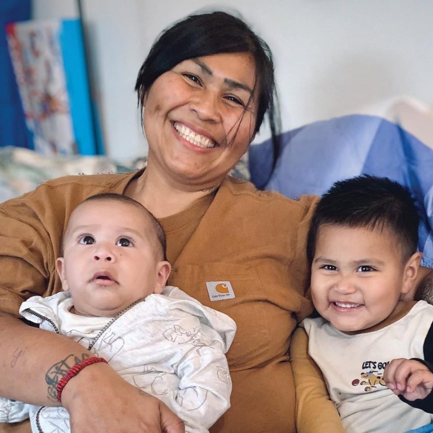 A woman smiling with two young children, one infant and one toddler, in a cozy indoor setting.
