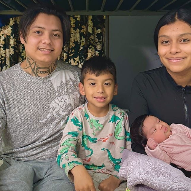 A family of four, including two adults, a young boy, and a baby, sit together on a bed under a bunk in a cozy room, finding safety and support at Mission amid family persecution.