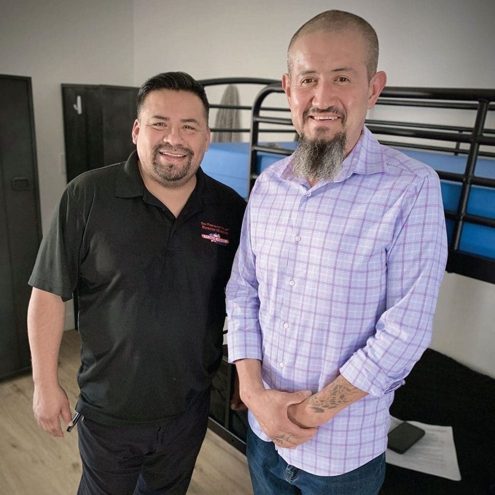 Rodolfo and his friend stand indoors near bunk beds, both smiling warmly. One sports a black shirt, the other a plaid one. A program lies open on the table covered with papers, hinting at their dedication and whole-hearted involvement in their shared passion.