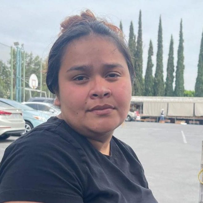 A woman with tied-back hair and a black shirt sits in a parking lot, surrounded by cars, trees, and a basketball hoop—embracing the challenge to survive day by day.