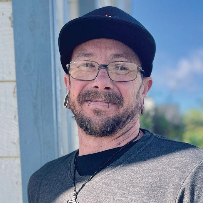 A compassionate man with tattoos and piercings, smiling outdoors in front of a building.