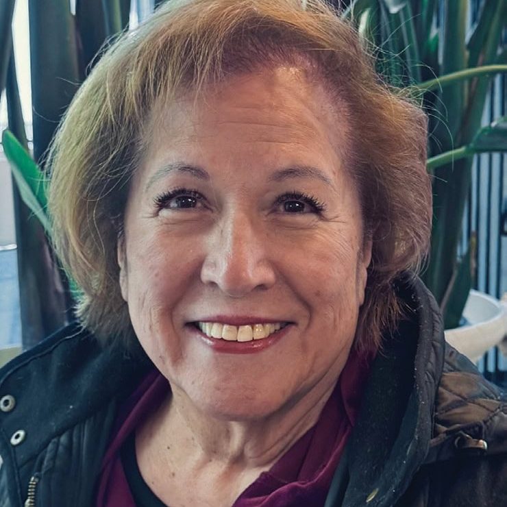 A smiling woman in a black jacket with greenery in the background.